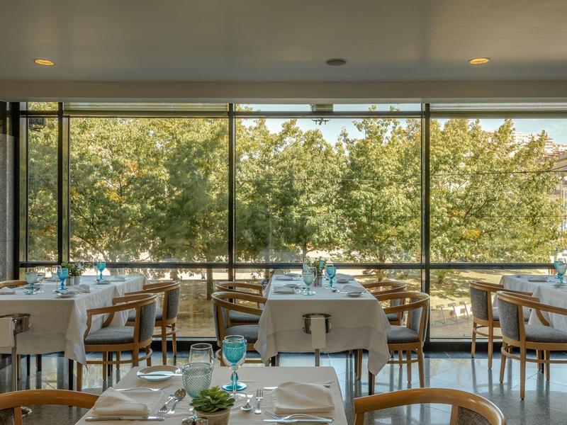Licht restaurant met houten stoelen, witte tafelkleden en grote ramen met uitzicht op groene bomen.