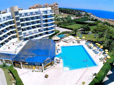 Hotelový komplex s velkým bazénem, slunečníky, zelenými plochami a výhledem na moře za jasné oblohy.