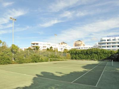 Freiluft-Tennisplatz mit grüner Umzäunung und Gebäuden im Hintergrund unter blauem Himmel