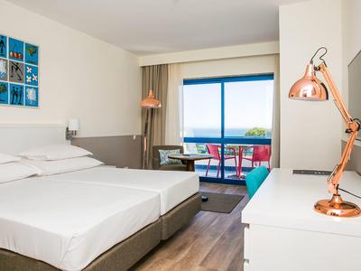 Světý hotelový pokoj se dvěma jednolůžky, stolem, balkonem s výhledem na moře a moderními lampami.