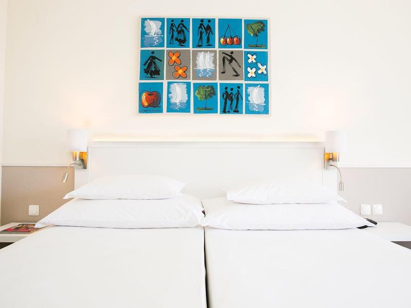 Dvě bílá jednolůžka s jednoduchým uměleckým dílem na stěně v světlém hotelovém pokoji.