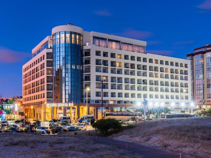 Edificio de hotel moderno de varios pisos por la noche con ventanas iluminadas y estacionamiento adelante.