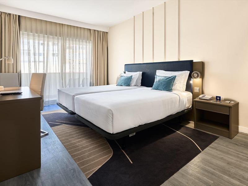 Habitación de hotel moderna con cama grande, escritorio y grandes ventanas con cortinas.