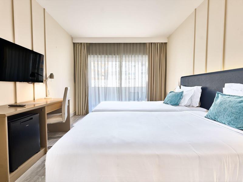 Habitación de hotel luminosa con dos camas grandes, TV, escritorio y ventana de piso a techo con cortina.