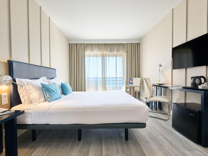 Habitación de hotel moderna con cama grande, televisor y vista al balcón y al mar.