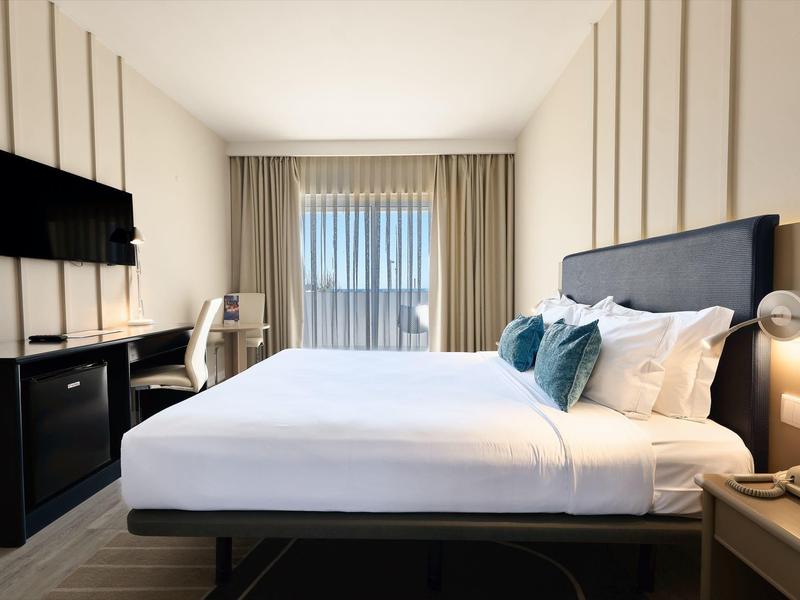 Habitación de hotel moderna con cama grande, TV, escritorio y ventana grande con cortinas.