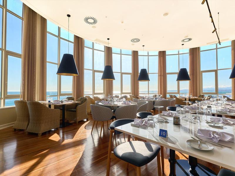 Restaurante luminoso con grandes ventanales, mesas, sillones y lámparas modernas con vistas al mar.