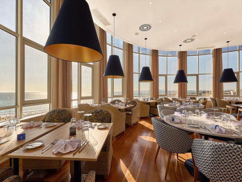 Restaurante elegante con grandes ventanales y vista al agua, tonos cálidos de madera e iluminación moderna.