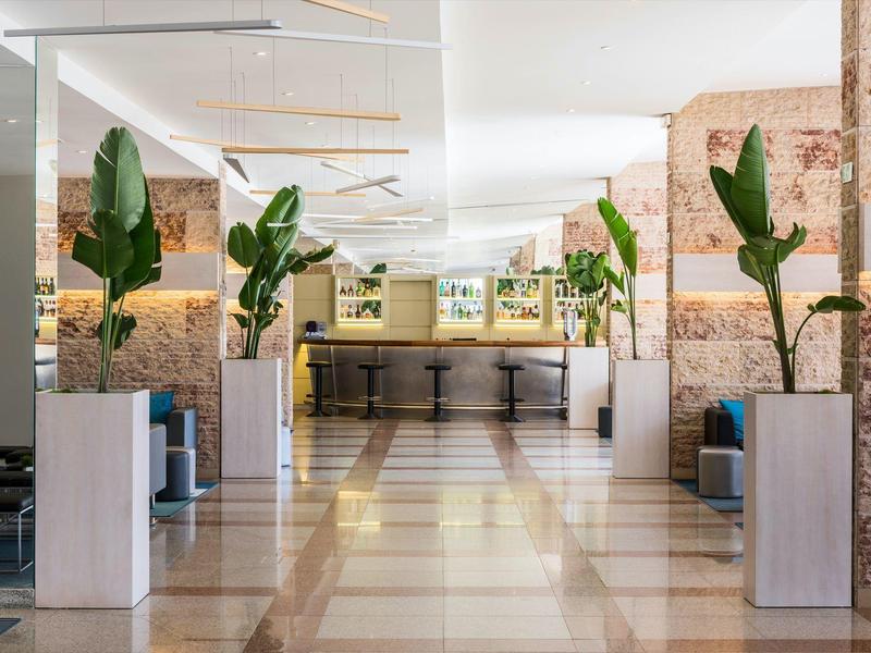 Luminoso vestíbulo de hotel con plantas, áreas de asiento y barra con taburetes al fondo.