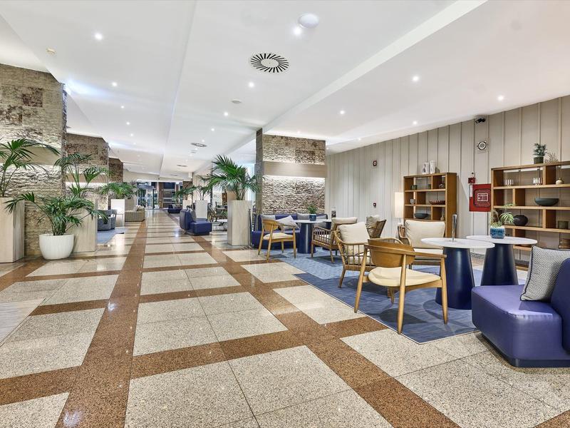 Amplio vestíbulo de hotel con asientos modernos y plantas decorativas junto a un piso de baldosas.