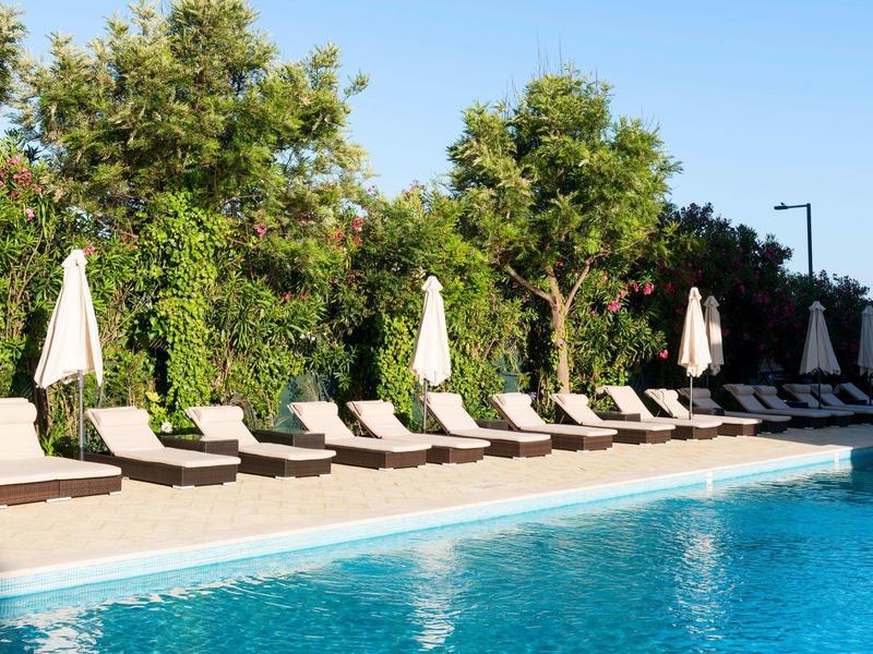 Piscina con agua azul y filas de tumbonas con sombrillas junto a árboles verdes.