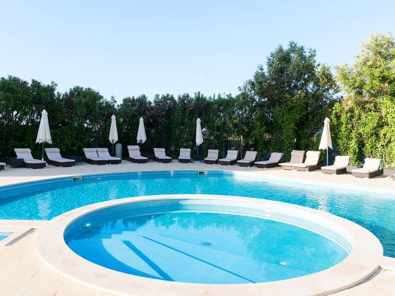 Piscina redonda con agua clara frente a una fila de tumbonas y sombrillas, rodeada de árboles.
