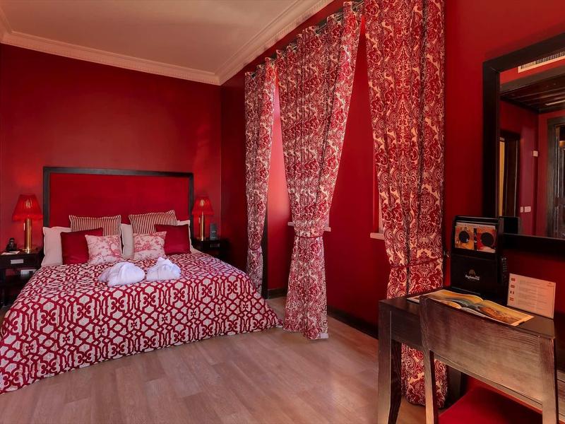 Camera da letto rossa con copriletto decorato, tende e mobili in legno.