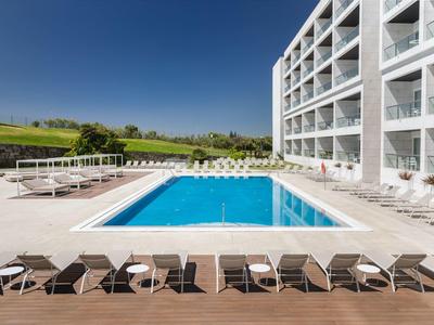 Modern outdoor pool with loungers and furniture next to a white hotel building under clear sky.