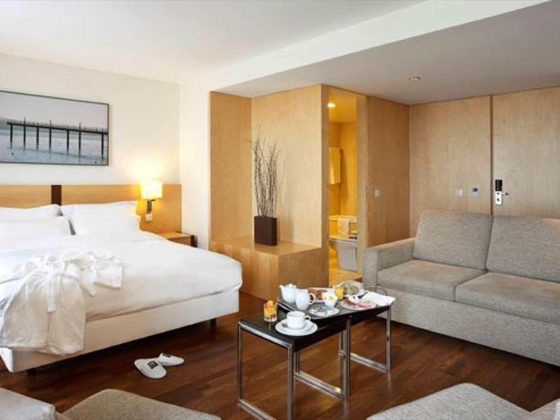 Modernes Hotelzimmer mit Doppelbett, grauem Sofa, Holzmöbeln und einem Kaffee-Tablett auf Tisch.