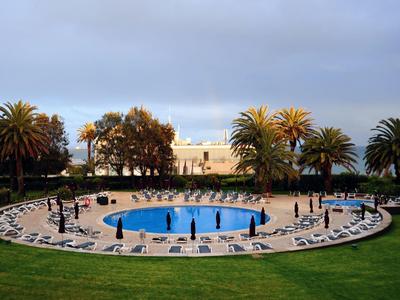 Round swimming pool surrounded by sun loungers, palm trees, and a white building under cloudy sky
