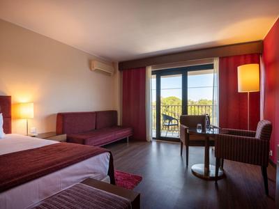 Hotel room with bed, sofa, armchair, table, and balcony overlooking greenery.