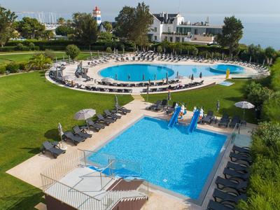 Two blue swimming pools with sun loungers and umbrellas in a green garden near a body of water.