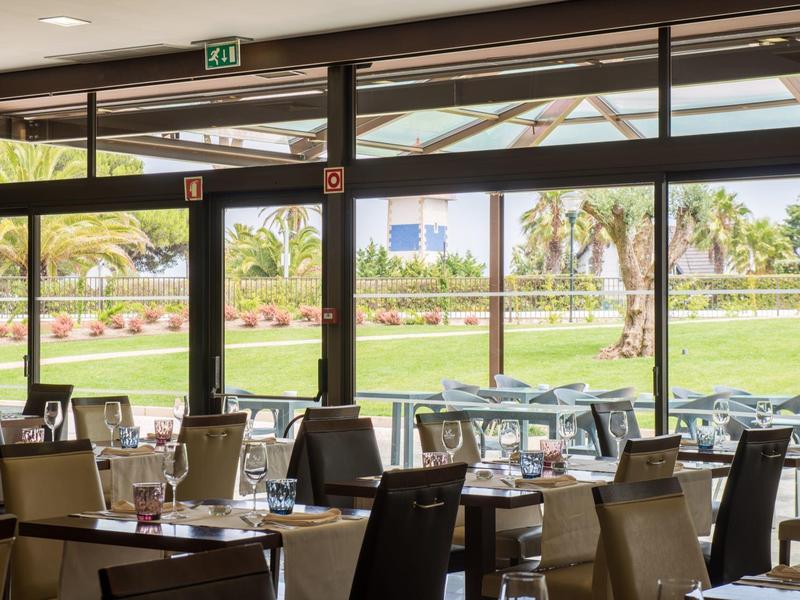 Hotel dining area with set tables and large windows overlooking a garden with palm trees.