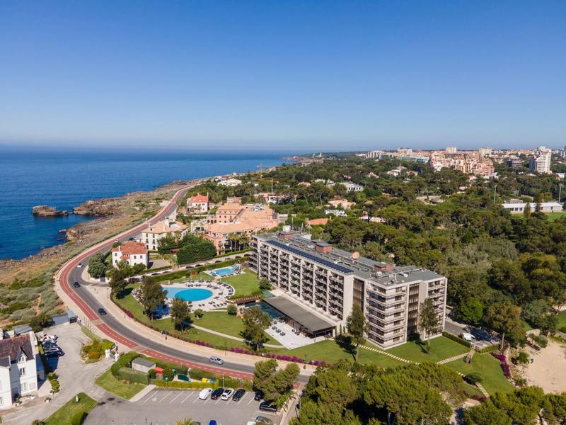Aerial view of a coastal hotel surrounded by greenery, roads, and a swimming pool under a clear blue sky.