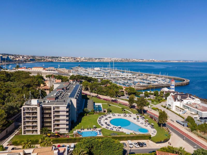 Aerial view of a coastal hotel with swimming pools, green lawns, and a marina in the background.