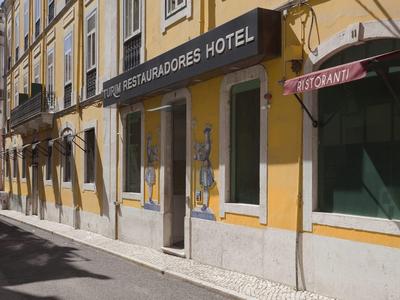 Enges Kopfsteinpflaster-Straße mit gelbem Hotelgebäude und grünen Fensterläden im Sonnenlicht.