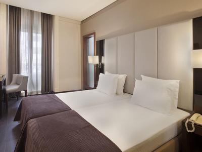 Modern hotel room with two single beds, desk, and natural light from a window.