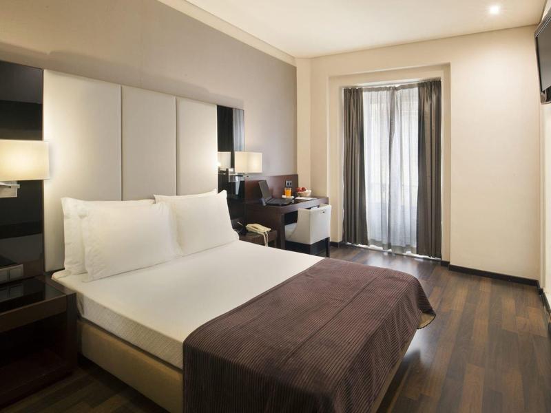 Bright hotel room with double bed, wooden floor, and balcony with curtains.