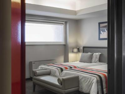 Schlichtes Schlafzimmer mit großem Fenster, grauem Bett, weißer Bettwäsche und gestreiftem Überwurf.