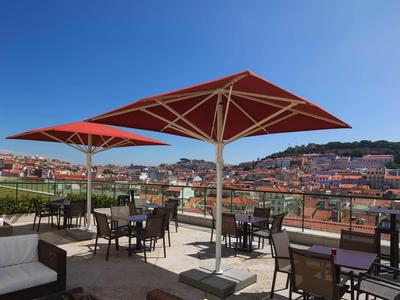 Dachterrasse mit roten Sonnenschirmen, Sitzgruppen und Blick auf Häuser und Hügel bei blauem Himmel.