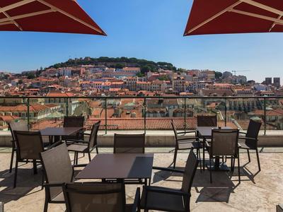 Offene Terrasse mit Tischen und Stühlen, Blick auf Stadt und Hügel unter blauem Himmel.