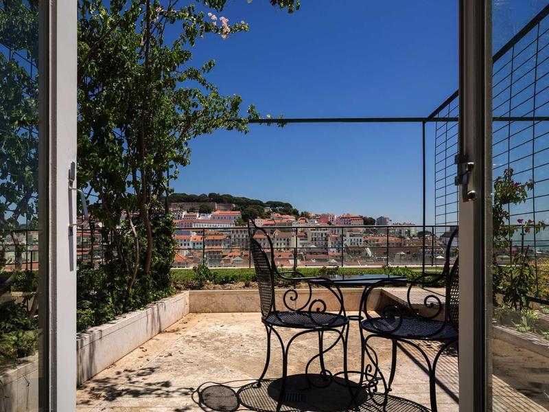 Blick durch offene Tür auf Balkon mit zwei schwarzen Metallstühlen und Tisch, im Hintergrund Stadt und blauer Himmel.