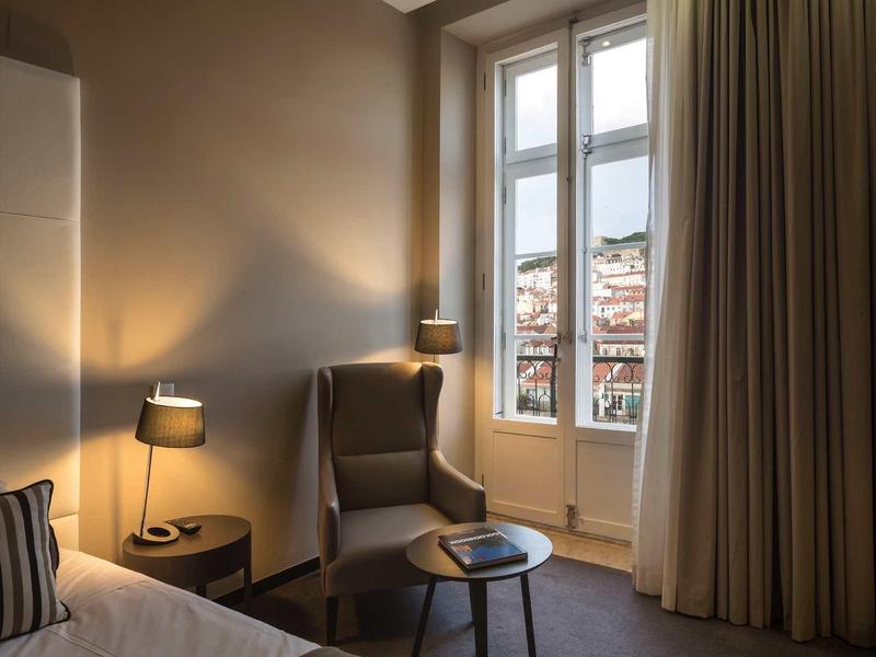 Gemütliches Schlafzimmer mit Sesseln, Nachttisch, Lampen und großen Fenstern mit Vorhängen.