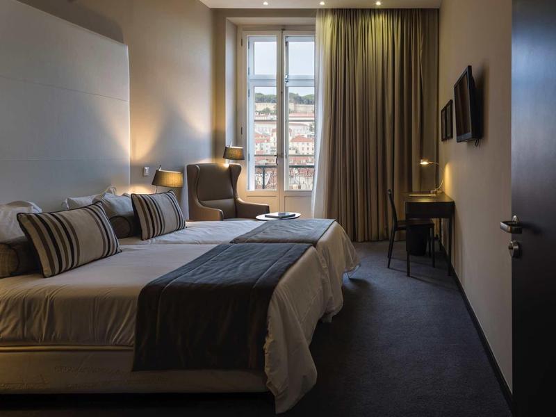 Modernes Hotelzimmer mit großem Bett, schlichten Möbeln und Fenster mit Vorhängen und Stadtblick.