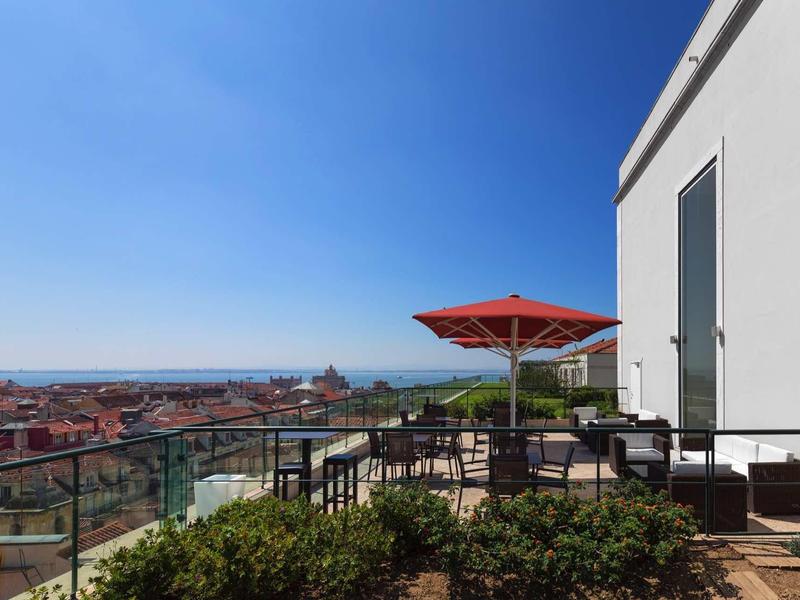 Dachterrasse mit Sitzbereich, rotem Sonnenschirm, Glasgeländer und Stadtblick unter klarem Himmel.