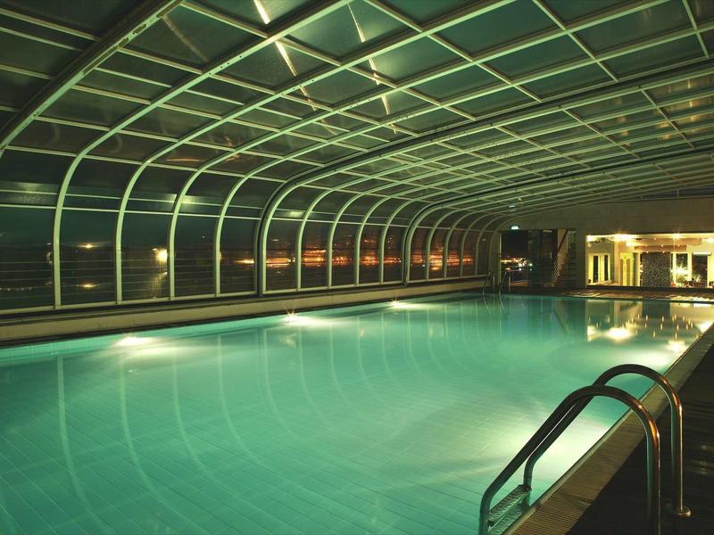 Verlicht binnenzwembad met transparant boogvormig dak en metalen reling.