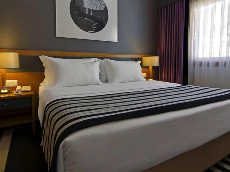 Moderne hotelkamer met tweepersoonsbed, twee nachtkastjes, lampen en raam met gordijnen.