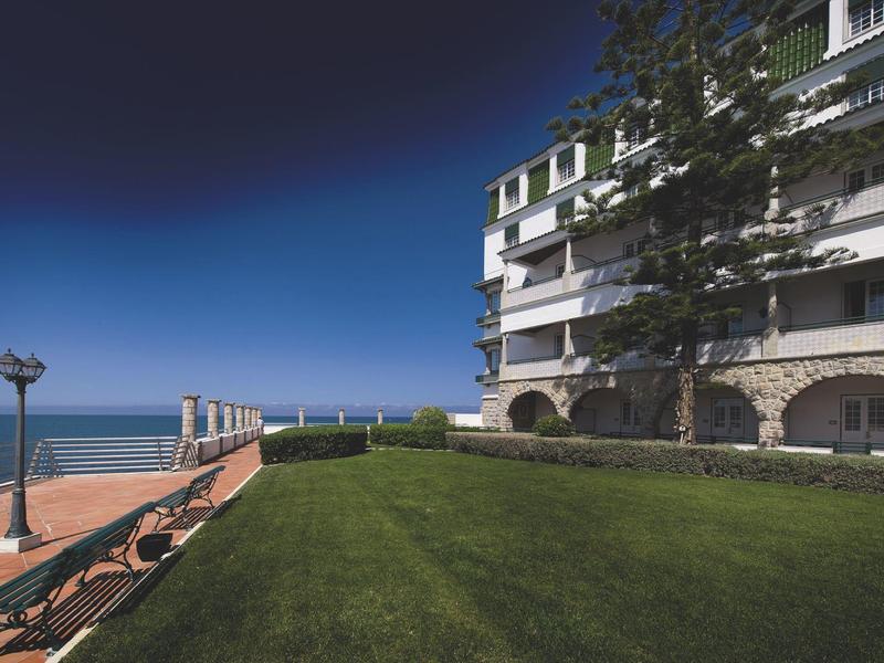 Zelený trávník před budovou u moře s lavičkou a lampou.