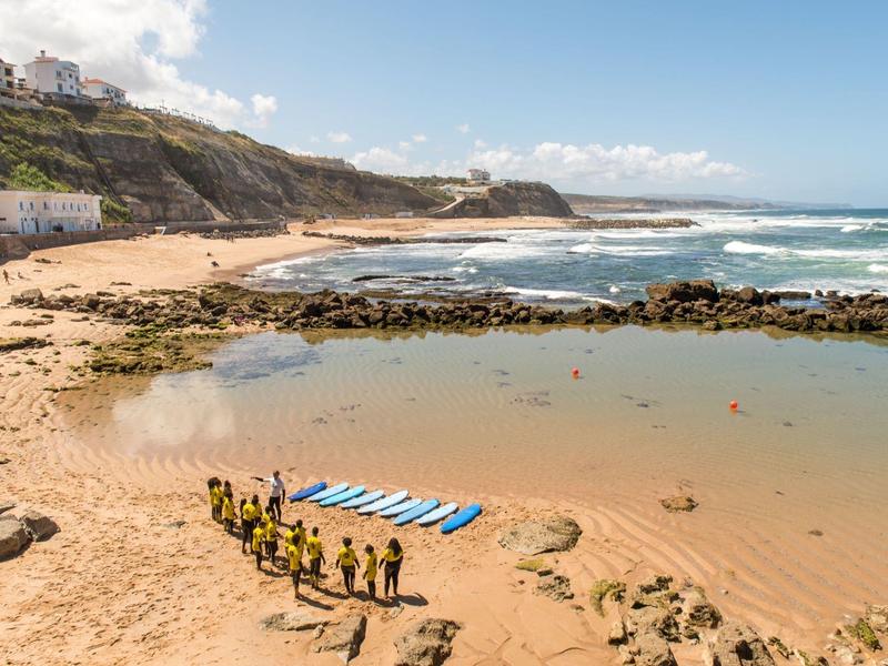 Pláž s skupinou lidí v žlutých oblecích a modrými deskami, skalnaté pobřeží v pozadí.