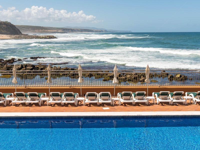 Bazén a lehátka před mořem s vlnami a částečně zataženou oblohou.