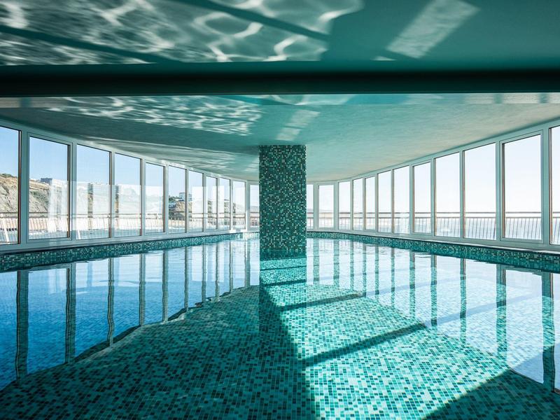 Vnitřní bazén s velkými okny s výhledem na moře a přirozeným světlem.