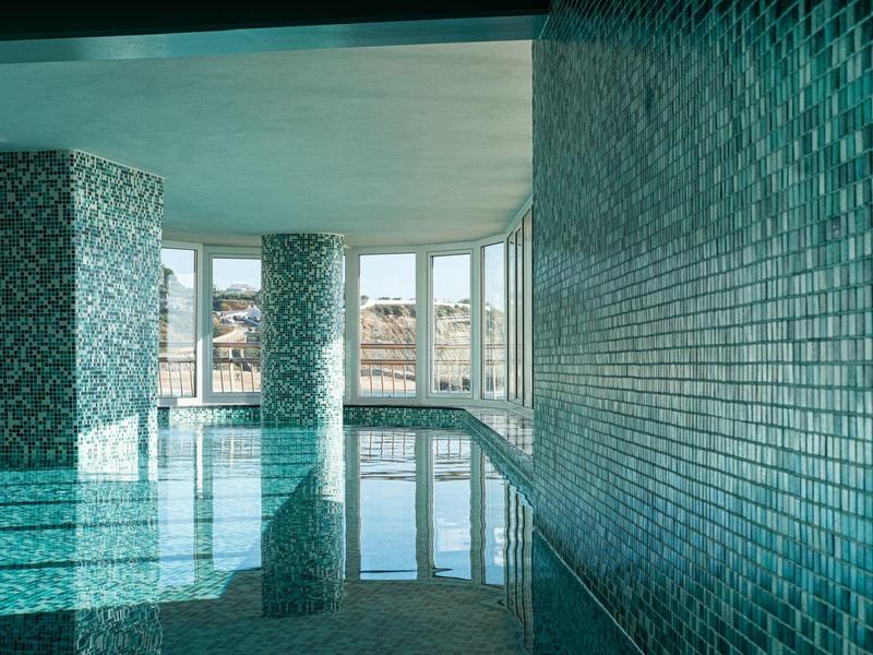 Moderní krytý bazén s modrými kachlovými stěnami a velkými panoramatickými okny.