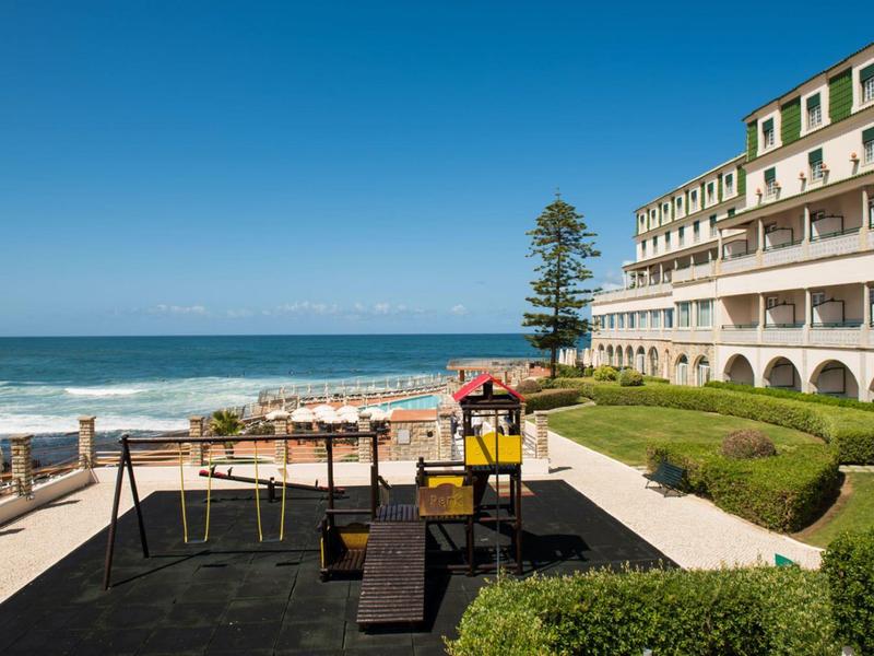Dětské hřiště s výhledem na oceán, hotel a zelený trávník pod jasnou oblohou.