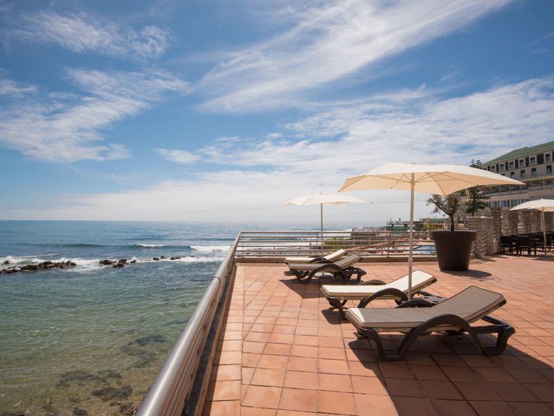 Slunná terasa s lehátky a slunečníky, výhled na klidné moře pod částečně oblačnou oblohou.