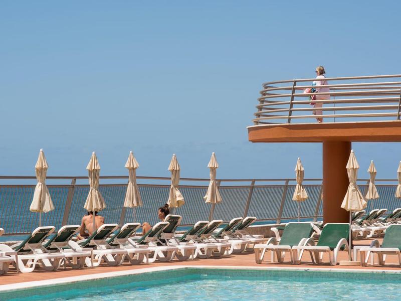 Hotelový bazén s lehátky, zavřenými slunečníky a výhledem na moře pod jasnou oblohou.