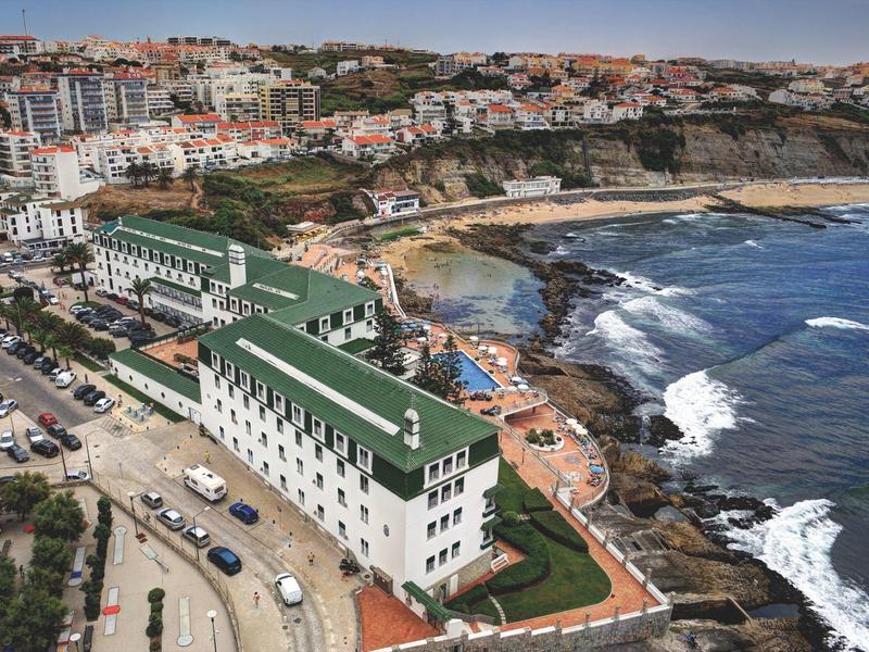 Pobřežní pohled na hotel se zelenými střechami u moře a kamenitou pláží, město v pozadí.