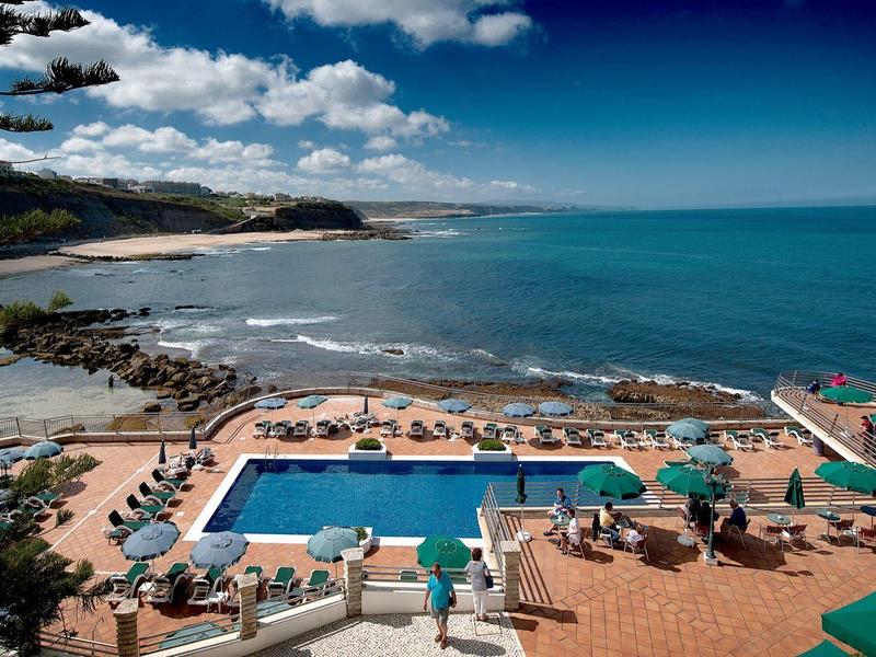 Bazén se zelenými slunečníky u kamenité pláže a modrého moře pod částečně zataženou oblohou.