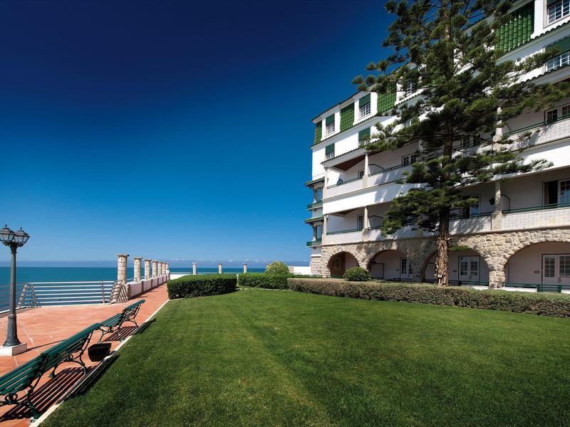 Zelený trávník vedle bílé budovy s výhledem na moře pod modrou oblohou.