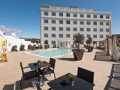 Piscina moderna de hotel con áreas de descanso y sombrillas frente a un edificio blanco de varios pisos.