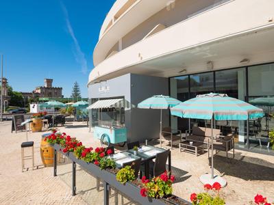 Terraza soleada de un hotel con sombrillas azules y flores rojas en macetas.